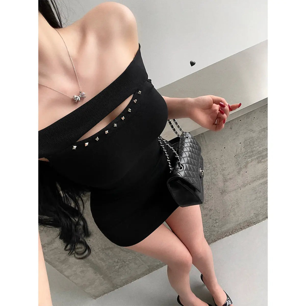 Close-up of a woman wearing a black ribbed off-shoulder mini dress with stud details, styled with a black quilted bag.