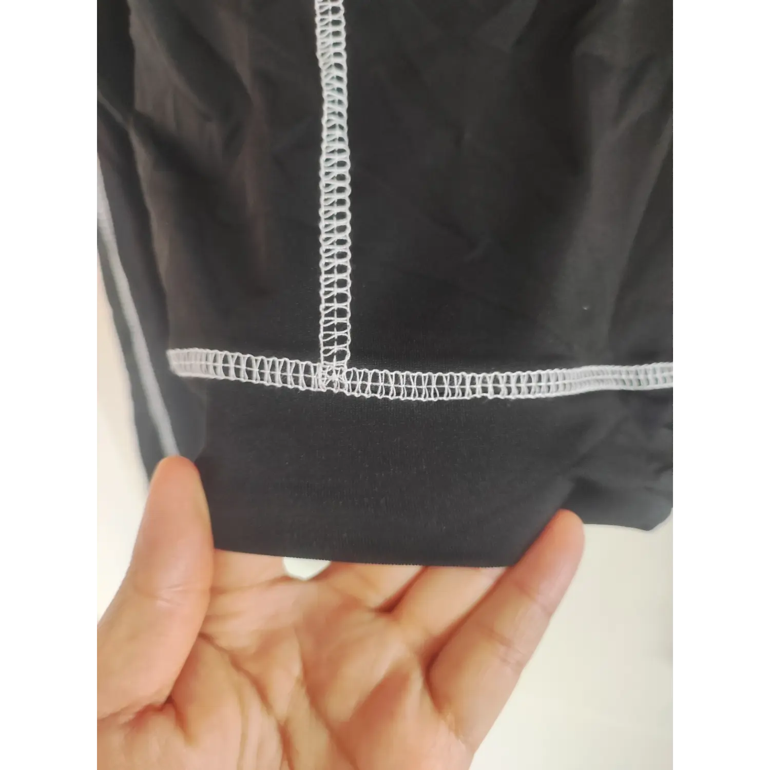Close-up photo of black stretch sportswear fabric with prominent white zigzag stitching, held in a human hand, showing elastic garment hem sewing detail
