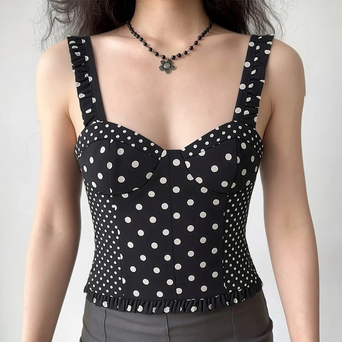 Model wearing a black and white polka dot bustier crop top with ruffled straps, styled with a black flower necklace and grey bottoms, front view.