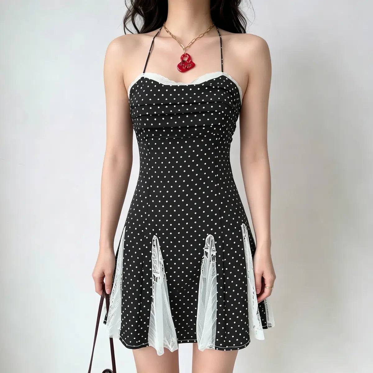 Model wearing a black and white polka dot halter-neck dress with lace panel details at the hem, carrying a brown handbag.
