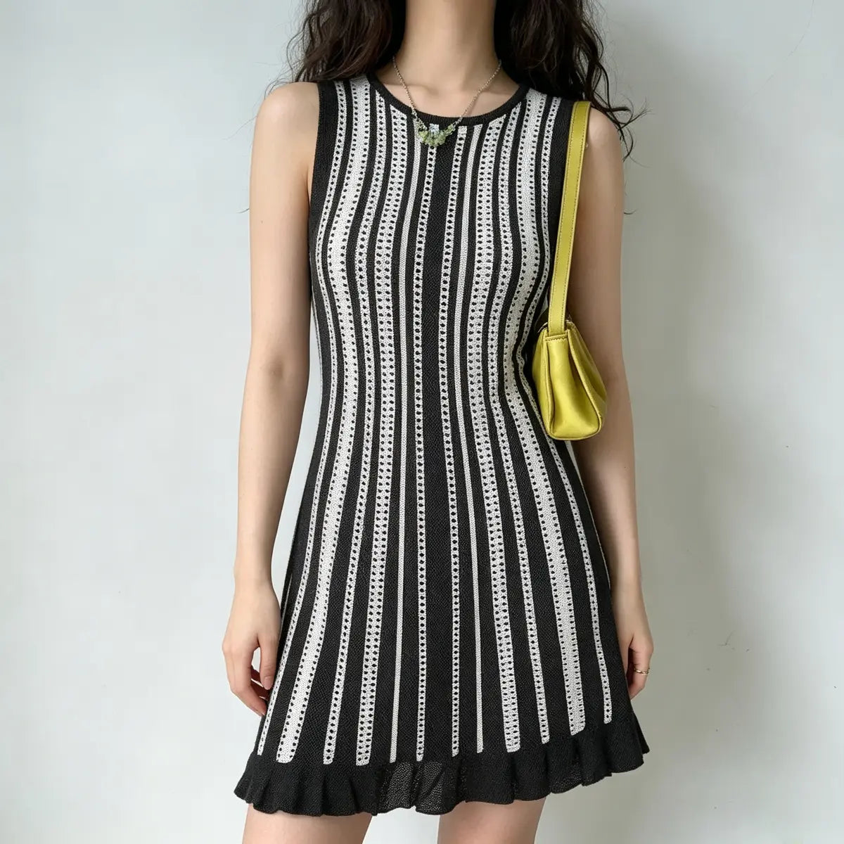 Model wearing a black and white striped knit mini dress with a ruffled hem, styled with a yellow handbag and green necklace, casual front pose.
