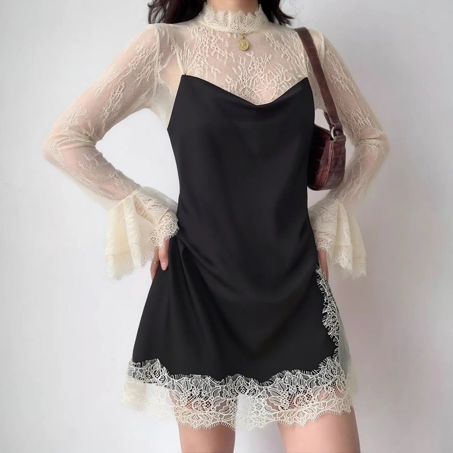 Model front view wearing black satin ruched slip mini dress with cream sheer lace mock‑neck bell‑sleeve overlay, eyelash lace hem