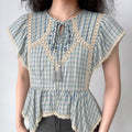 Front lifestyle shot on model of blue check cottagecore blouse, keyhole tassel neck, lace splicing, flutter sleeves, irregular peplum hem, vintage farm style top