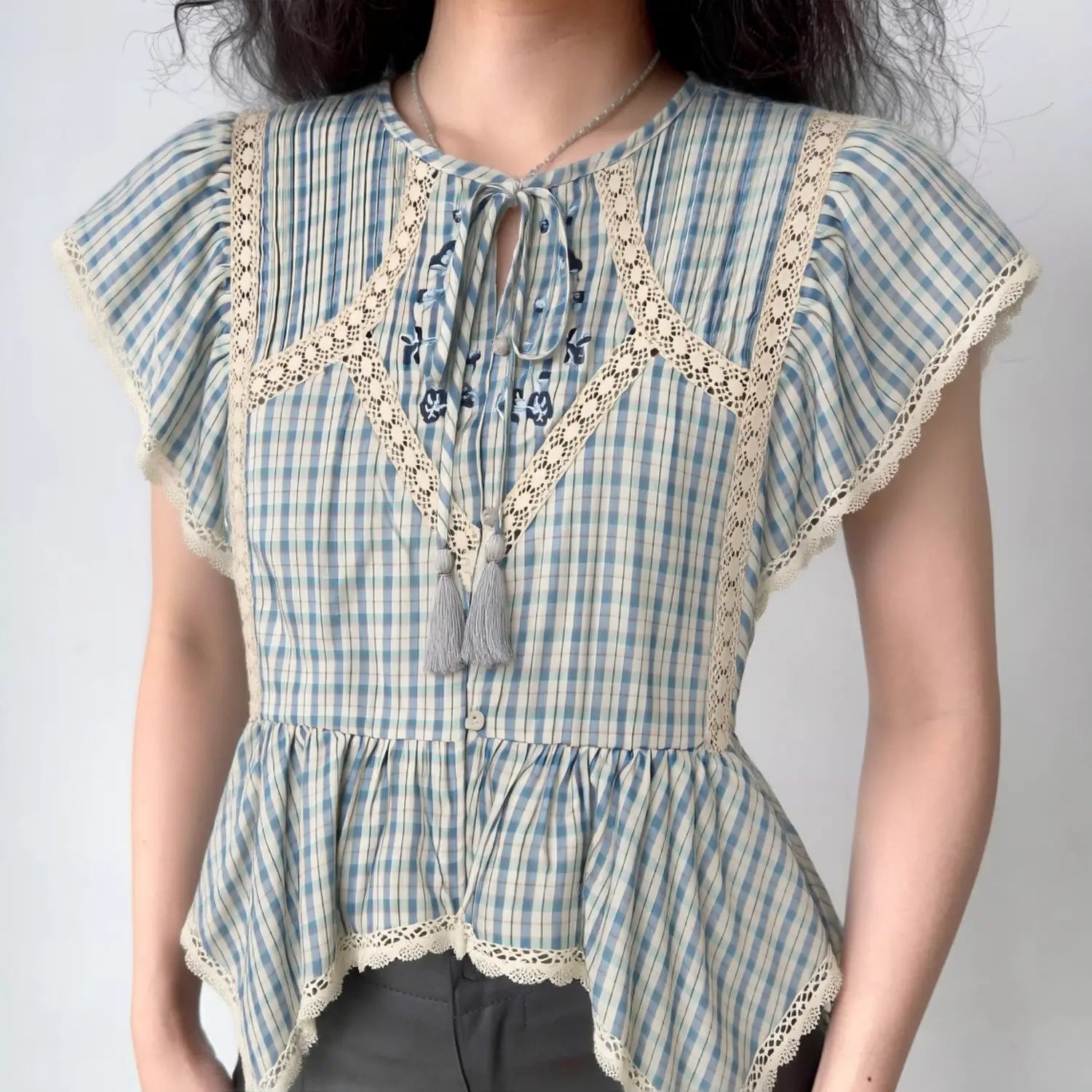 Front lifestyle shot on model of blue check cottagecore blouse, keyhole tassel neck, lace splicing, flutter sleeves, irregular peplum hem, vintage farm style top