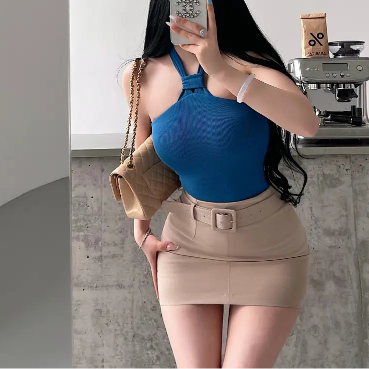 A woman wearing a blue ribbed keyhole halter top, styled with a beige belted mini skirt, taking a mirror selfie.