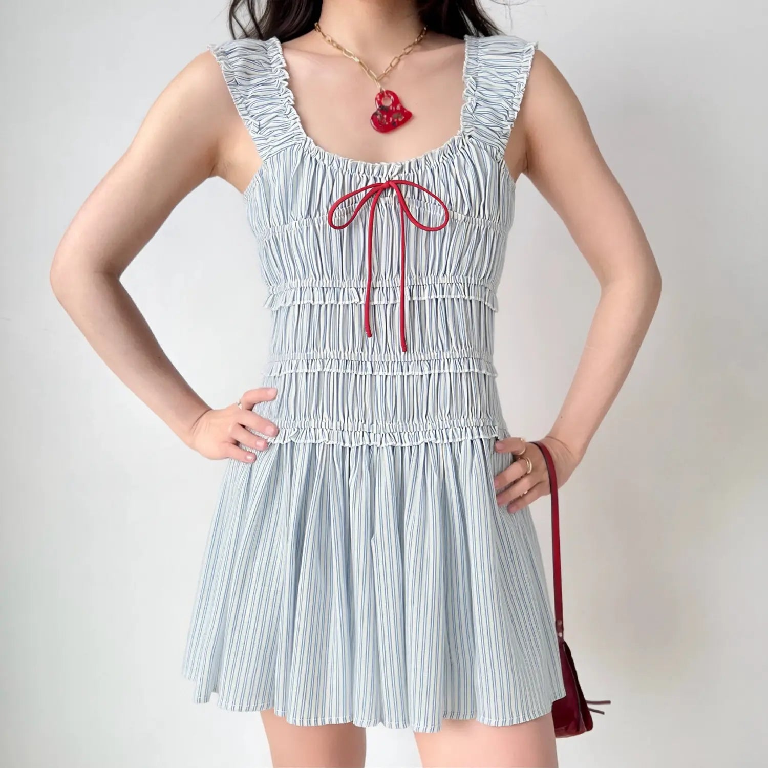 Model wearing blue‑white vertical stripe smocked mini dress, square neck, ruffle straps, contrast red front bow tie, flare short skirt, studio background
