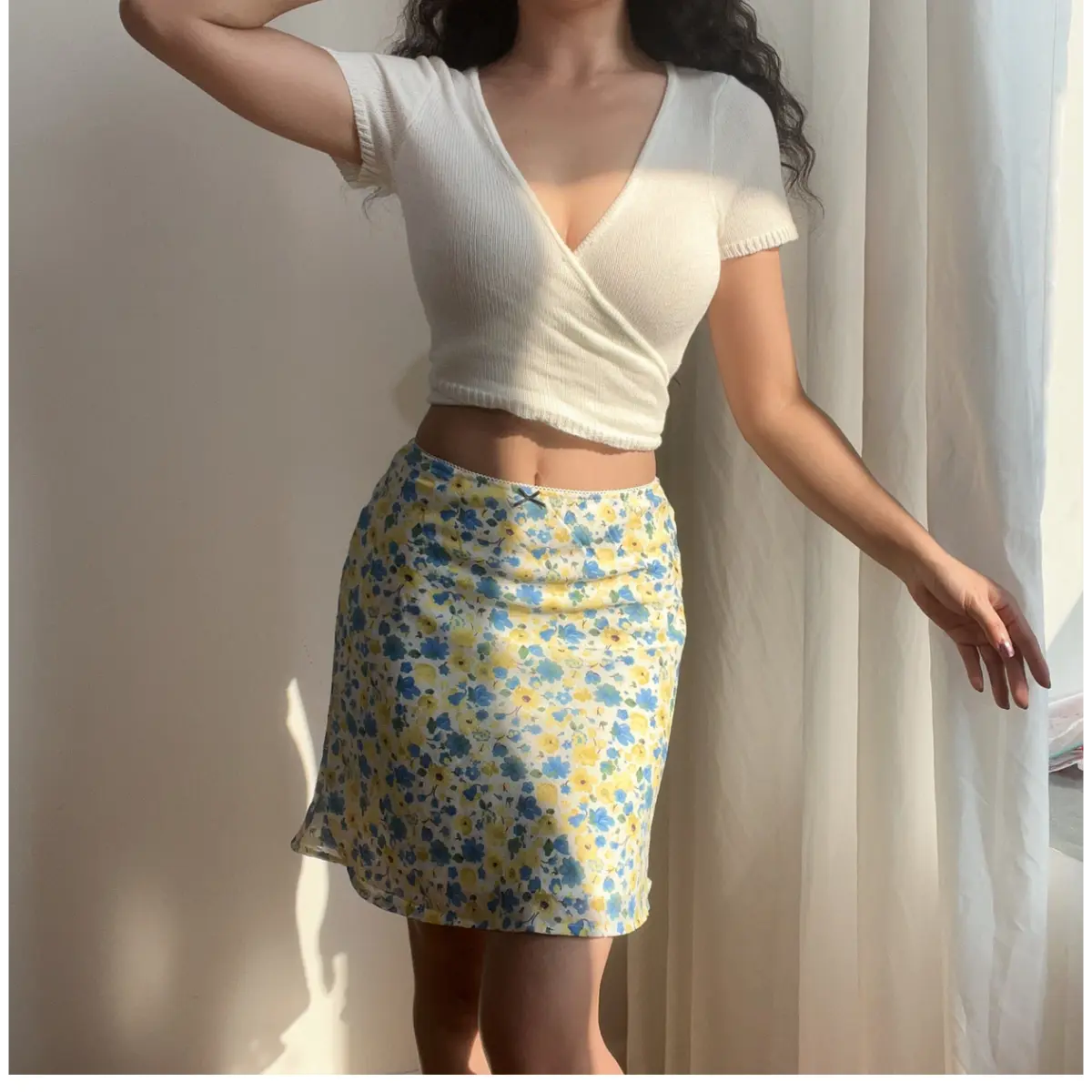 Side view of a woman wearing a blue and yellow floral chiffon mini skirt, paired with a white cropped knit top, showing the skirt's drape and length.
