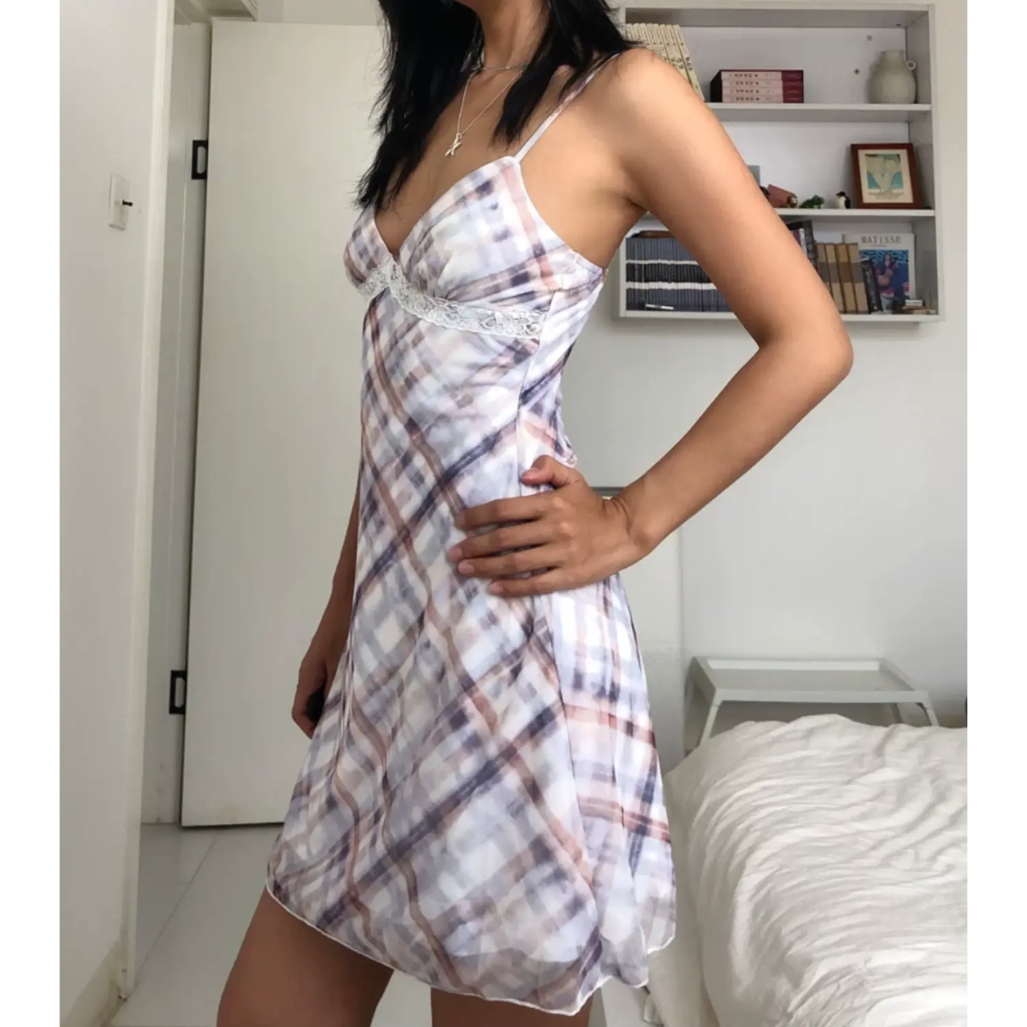 Medium full indoor bedroom lifestyle shot of sheer slip mini dress, model resting one hand on hip to highlight fit, adjustable thin plaid spaghetti straps, A-line ruffled mini hem