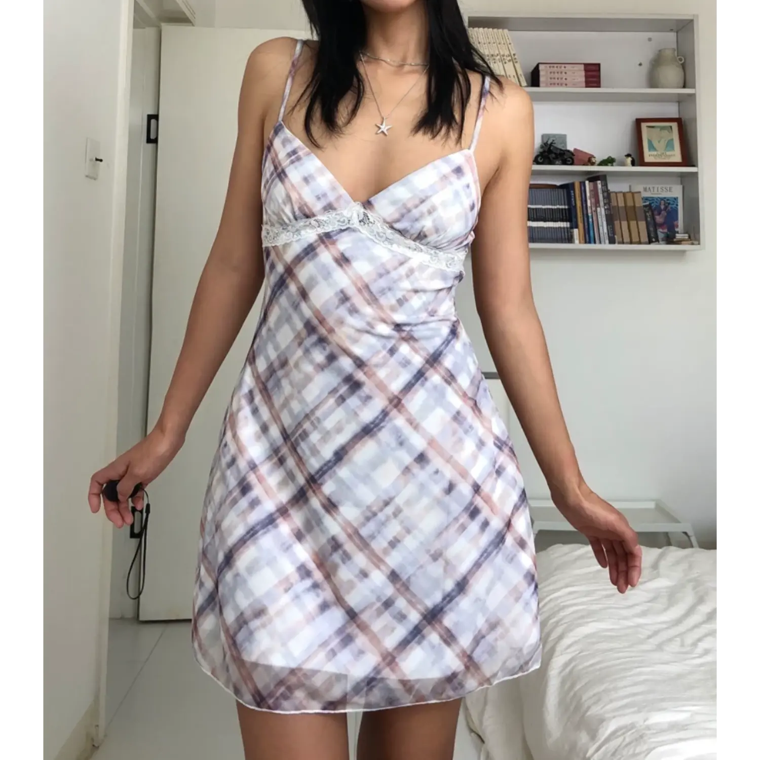 Cozy indoor bedroom casual full shot of watercolor plaid sheer mesh mini slip dress, white scalloped lace underbust detail, thin adjustable plaid straps, model spreading arms naturally, soft A-line ruffled mini hem, clean minimalist white home background with storage shelf.