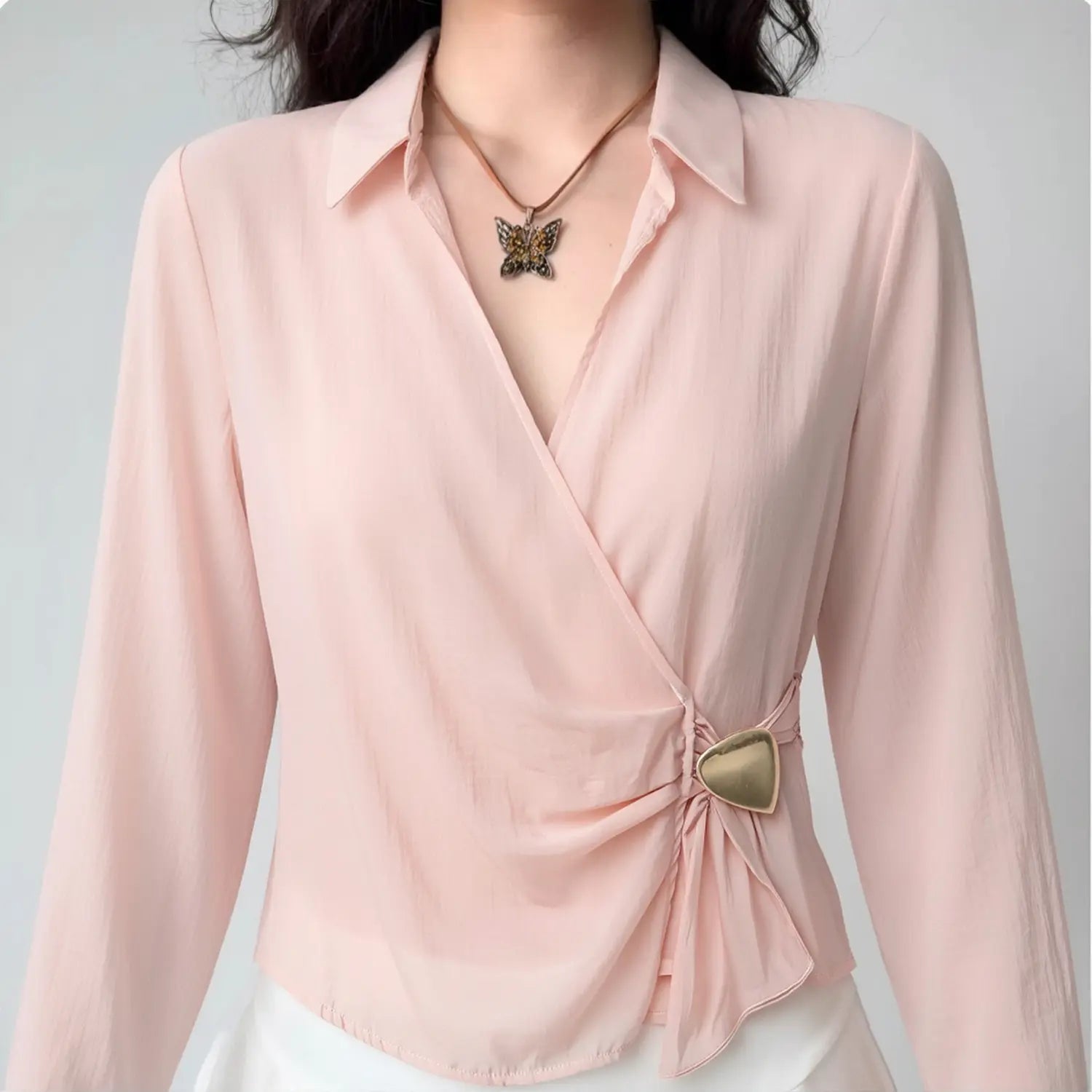 Front close lifestyle shot on model, pointed lapel wrap cross V-neck satin blouse, side waist fabric ruching fixed with irregular heart gold metal buckle, lightweight sheer blush satin fabric, collocated with butterfly pendant necklace and white bottoms, gentle elegant daily matching style