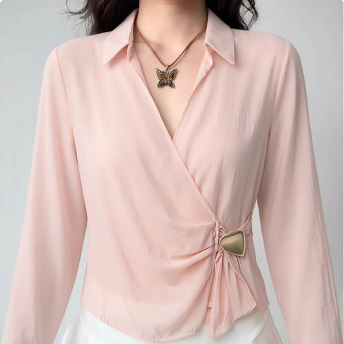 Front close lifestyle shot on model, pointed lapel wrap cross V-neck satin blouse, side waist fabric ruching fixed with irregular heart gold metal buckle, lightweight sheer blush satin fabric, collocated with butterfly pendant necklace and white bottoms, gentle elegant daily matching style
