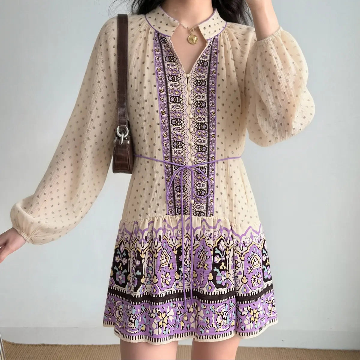 Woman styled in a boho printed mini dress with balloon sleeves, decorative front panel, and gathered hem, accessorized with a brown shoulder bag.