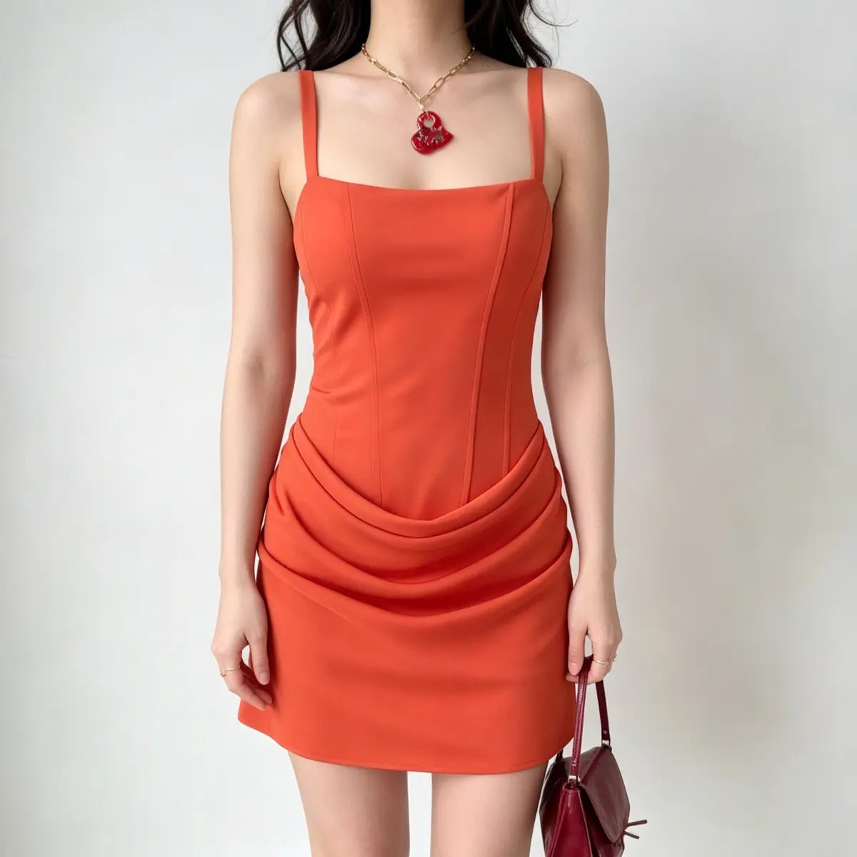 Model wearing a bright orange corset-style mini dress with a draped front detail and wide straps, carrying a red handbag.