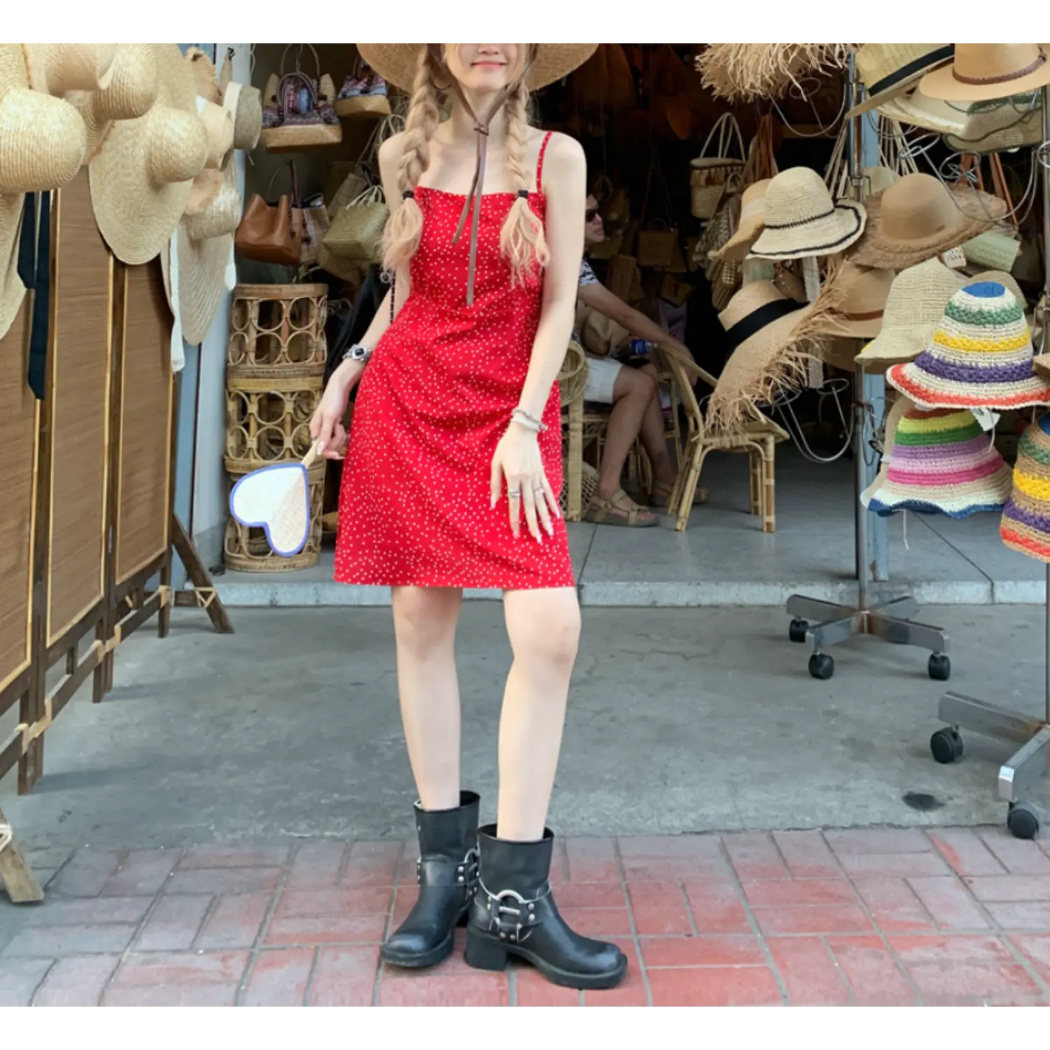 Full body travel street shot of red white polka dot mini slip dress, model wearing wide brim straw hat and silver buckle black ankle boots, background lined with rattan hats and woven furniture displays