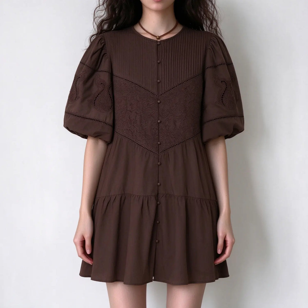 Model wearing brown embroidered mini dress with puff short sleeves, front button placket, eyelet lace embroidery, tiered hem, white background
