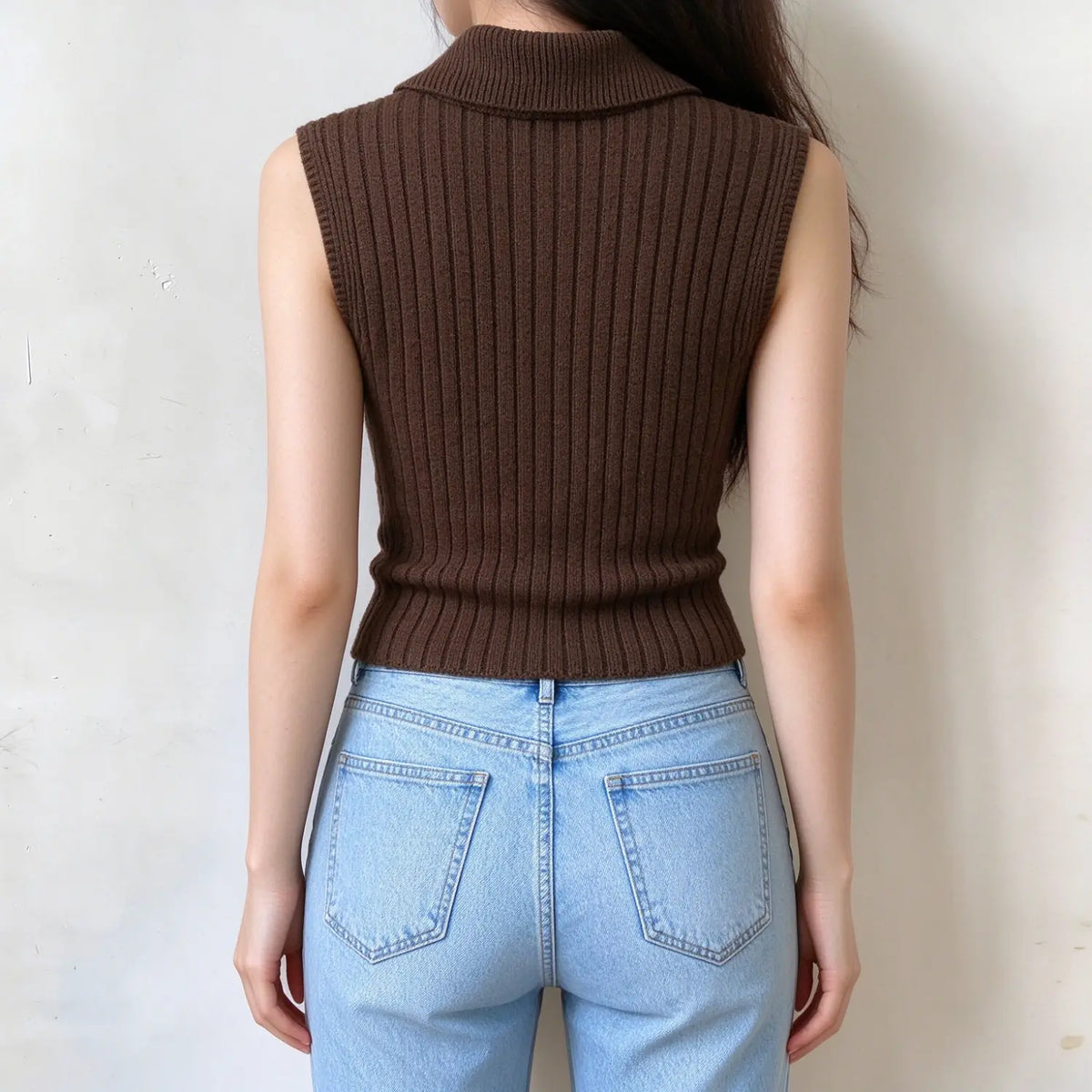 Back view of model in brown ribbed knit sleeveless zip vest, vertical rib texture, cropped fit, matched with light blue denim jeans