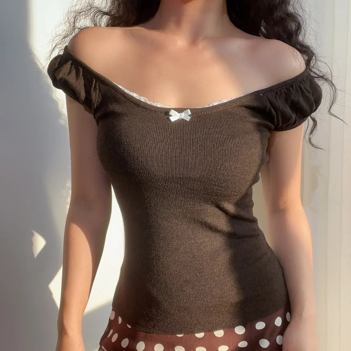 Woman wearing a brown ribbed off-the-shoulder top with puff sleeves, lace trim, and a small white bow, paired with a brown and white polka dot skirt.