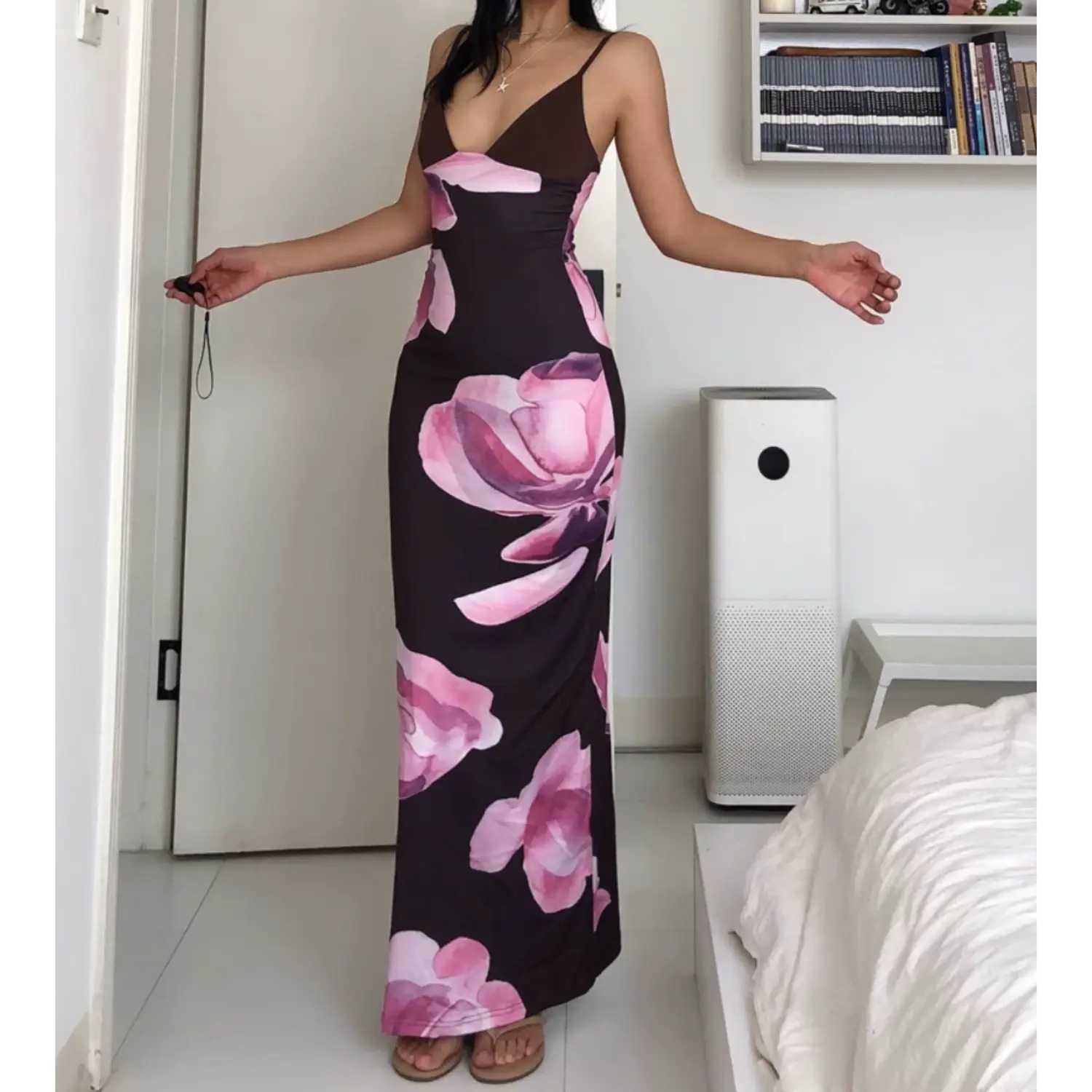 Full body indoor bedroom lifestyle shot of slim fit slip maxi dress, model spreading arms casually, wearing flat slide sandals, dark brown base with oversized gradient pink magnolia floral print