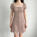  Model wearing a brown and white gingham mini dress with lace trim at the neckline and sleeves, a buttoned front, and patch pockets.

