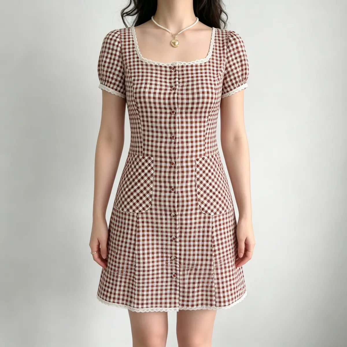  Model wearing a brown and white gingham mini dress with lace trim at the neckline and sleeves, a buttoned front, and patch pockets.
