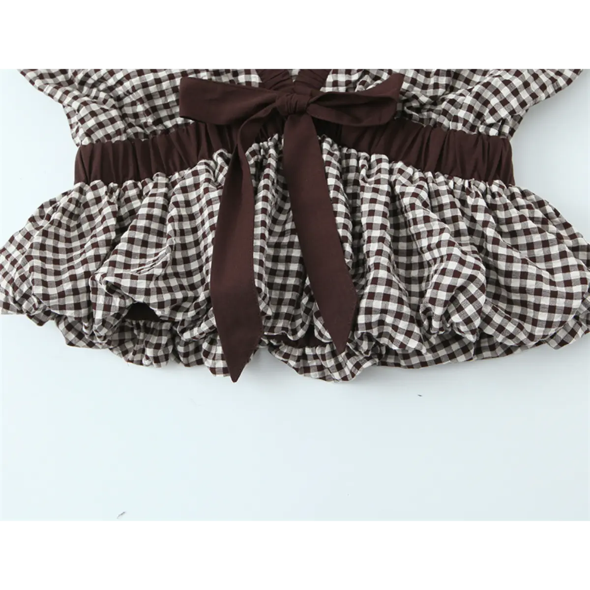Detail view of the waist on the gingham top, showing the brown fabric bow, elasticated waistband, and ruffled hem
