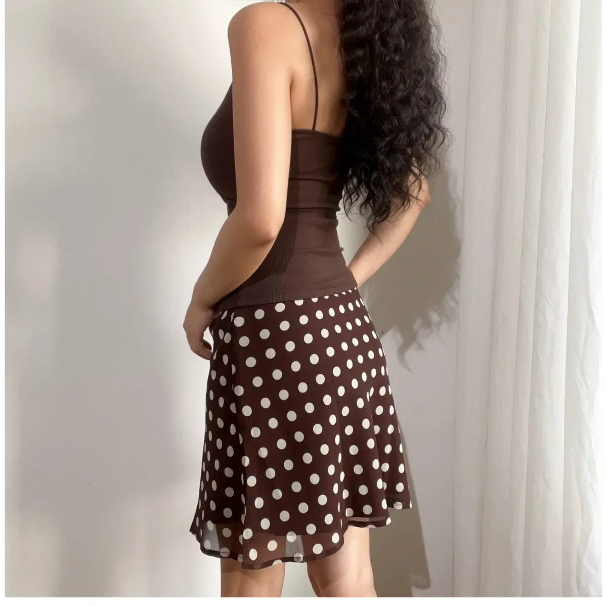  Side view of a woman wearing a brown and white polka dot chiffon mini skirt, paired with a brown ribbed camisole, showing the skirt's drape and length.