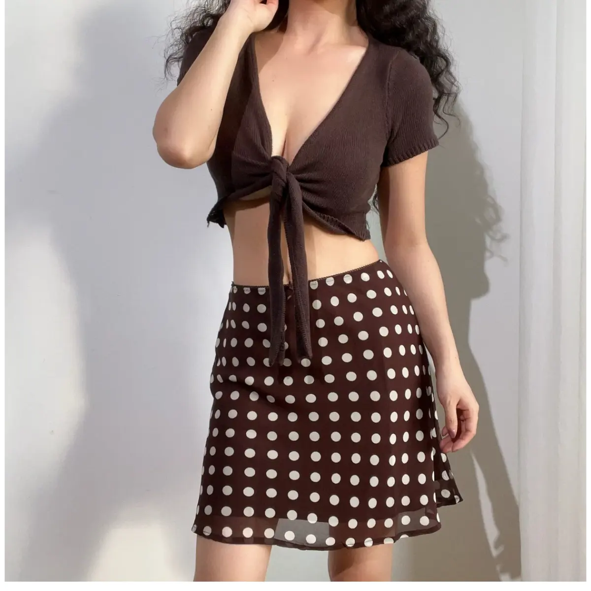 Woman wearing a brown and white polka dot chiffon mini skirt, paired with a brown tied-front knit cardigan, showing the skirt's silhouette.