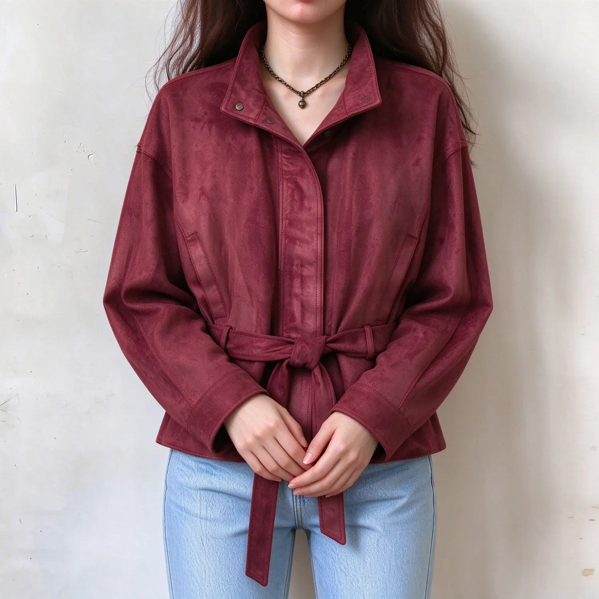 Female model wearing burgundy suede look belted outerwear, stand collar, tied waist sash, hands clasped in front to show tailored silhouette