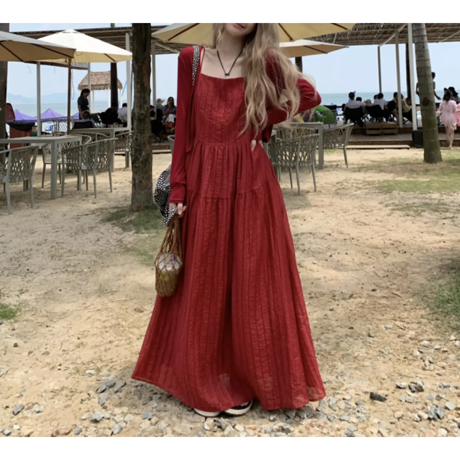 Coastal beach outdoor lifestyle shot of burgundy textured tier slip maxi dress, layered sheer red long sleeve cover up, model carrying woven rattan mesh bag, slide sandals on gravel sand