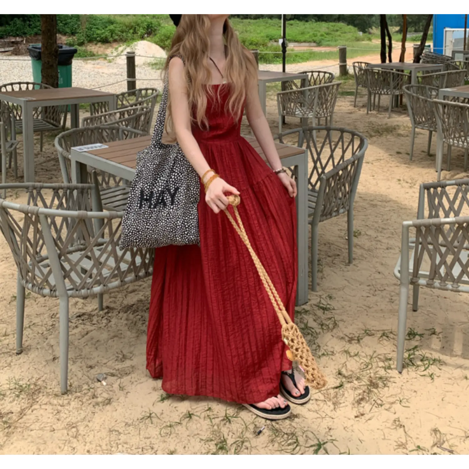Seaside outdoor sitting lifestyle shot of rich red vertical textured tier maxi dress, model seated beside grey rattan patio furniture, holding woven braided straw bag strap, slide sandals visible