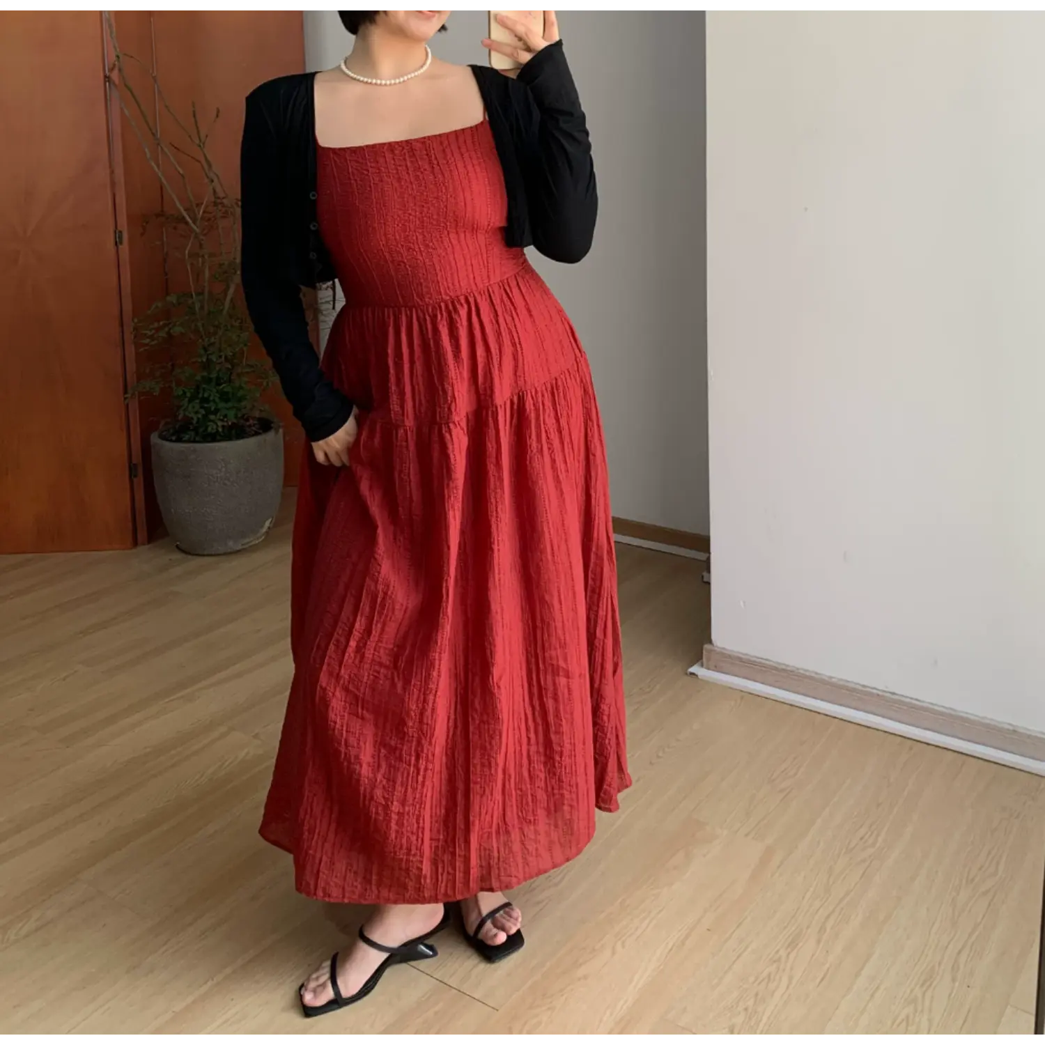 Medium full indoor mirror lifestyle shot of rich red vertical textured tiered maxi dress, layered black fitted cropped knit cardigan, model resting one hand on hip to show waist silhouette