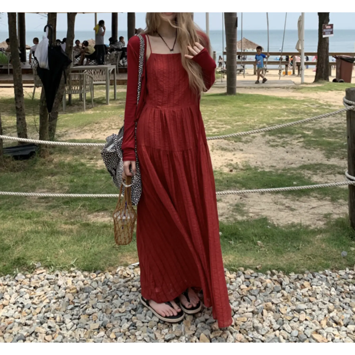 Vacation beach outdoor three-quarter shot of deep red crinkled tier maxi slip dress, thin red long sleeve layering cover up, woven rattan bag and black slide sandals on stone gravel path