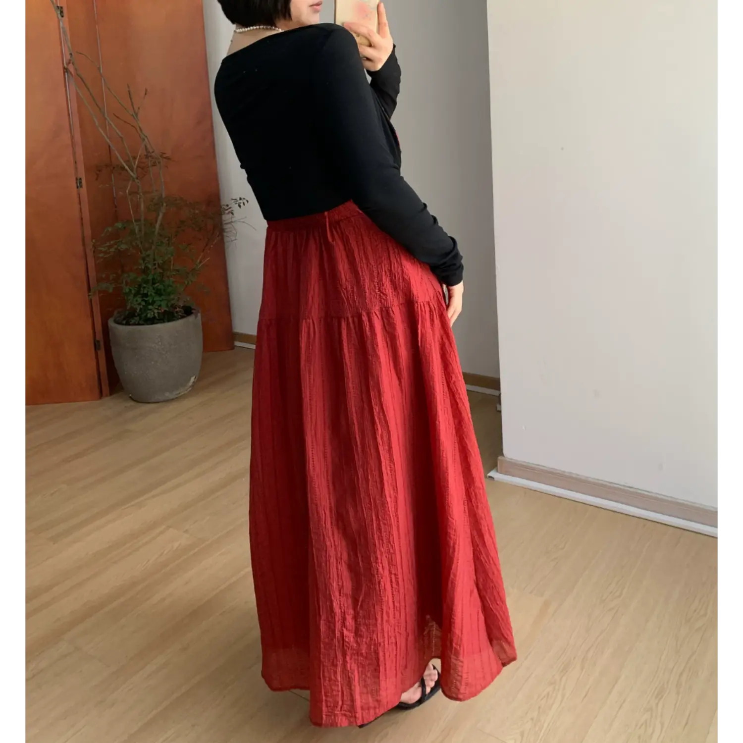 Full three-quarter back indoor mirror shot of deep red crinkled tier maxi slip dress, layered black cropped knit cardigan, showing long flowy layered skirt hem and rear tie detail