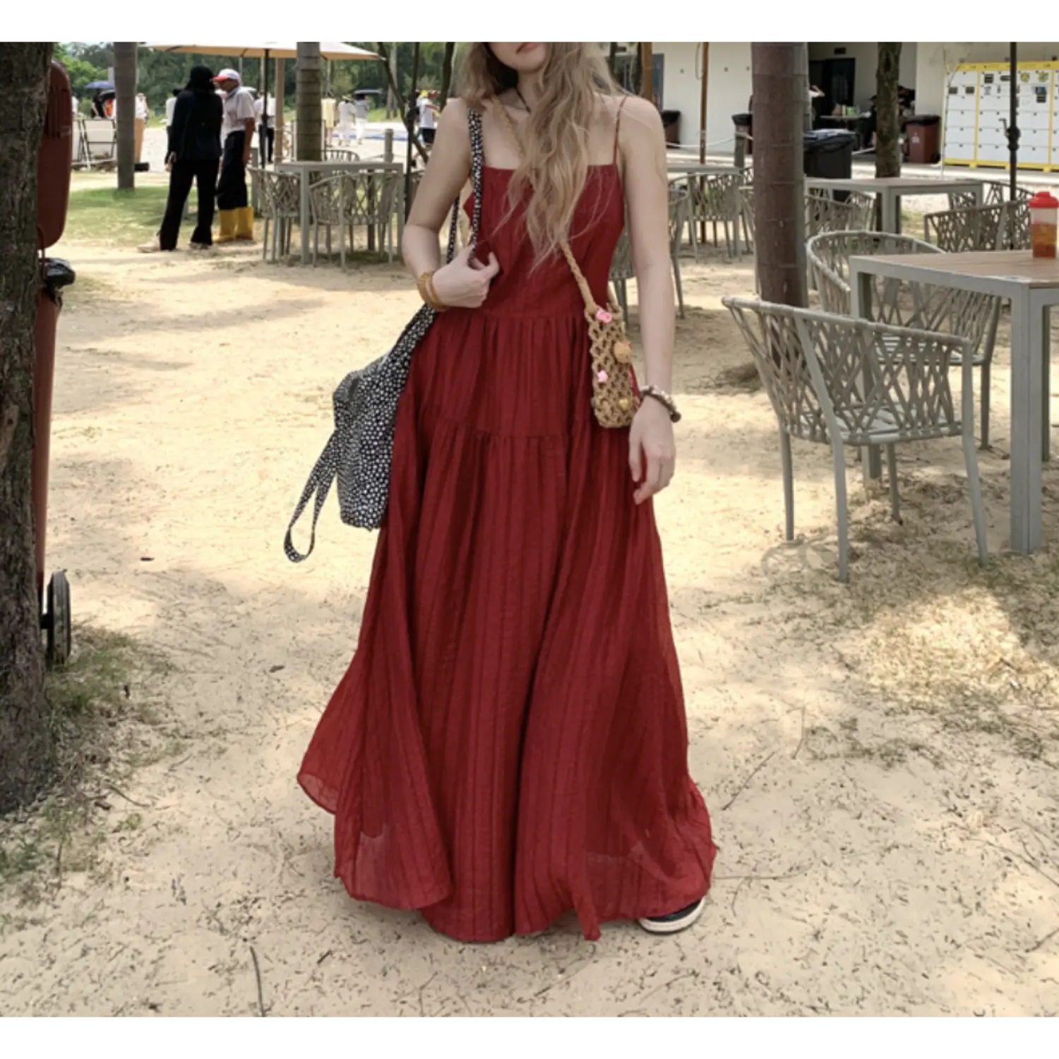 Full body coastal outdoor street shot of burgundy crinkled tier slip maxi dress, sheer red long sleeve cover up layered over top, polka dot canvas crossbody bag and woven rattan mini pouch accessory