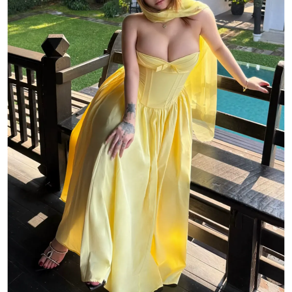 Woman in a butter yellow strapless bow dress on a wooden balcony, leaning against a railing with the pool in the background.