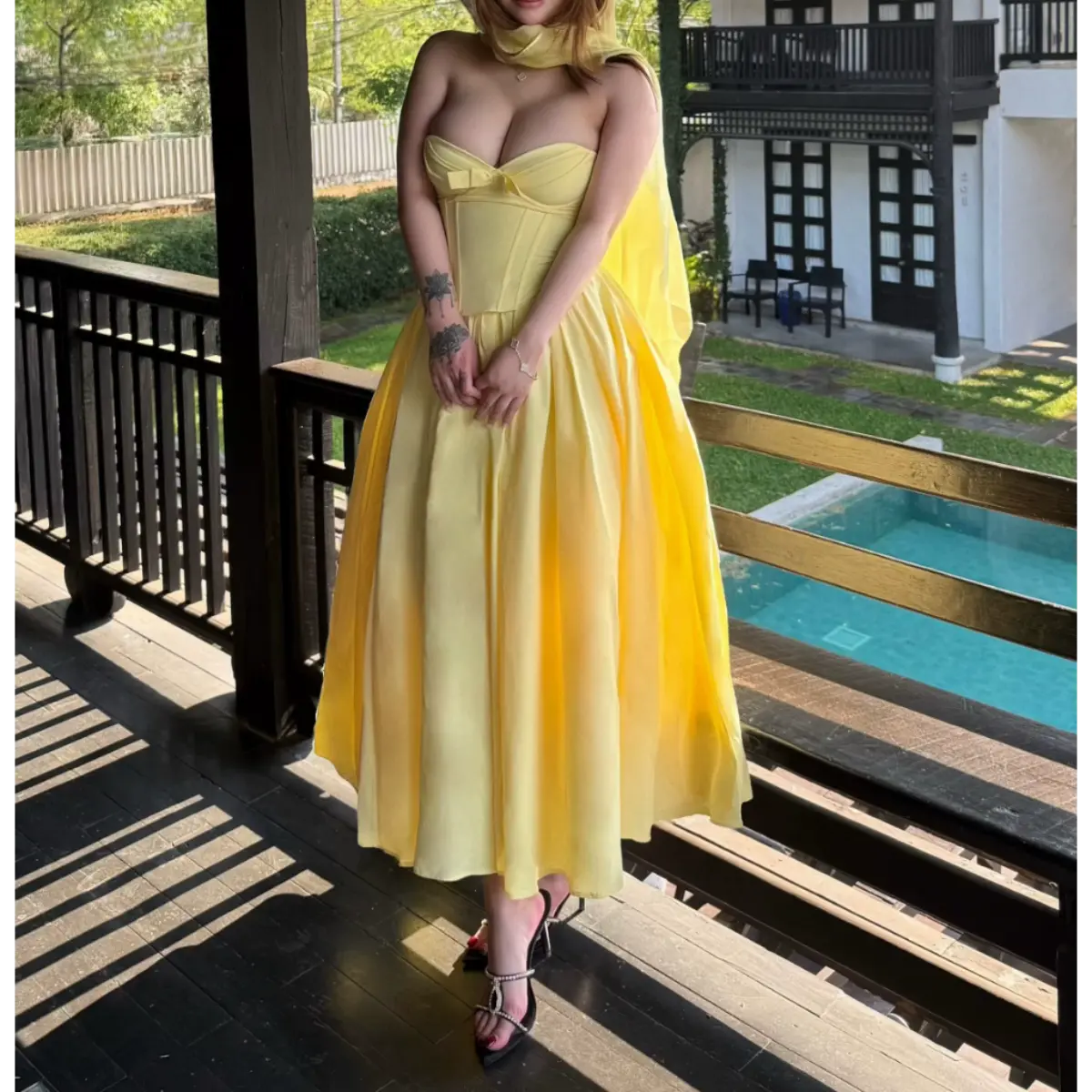 Woman in a butter yellow strapless bow-front dress by a pool, styled with a matching sheer shawl and black heels.