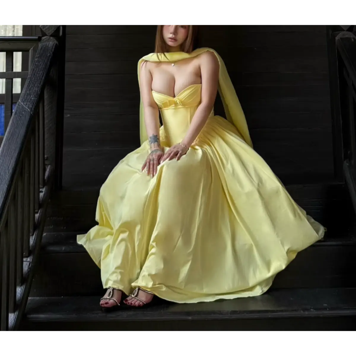Woman in a butter yellow strapless bow dress seated on dark wooden stairs, showcasing the dress’s voluminous skirt and matching shawl.