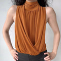 Front lifestyle shot on model of caramel glossy satin halter top, ruched high neck, front cross wrap drape, elegant sleeveless evening casual top