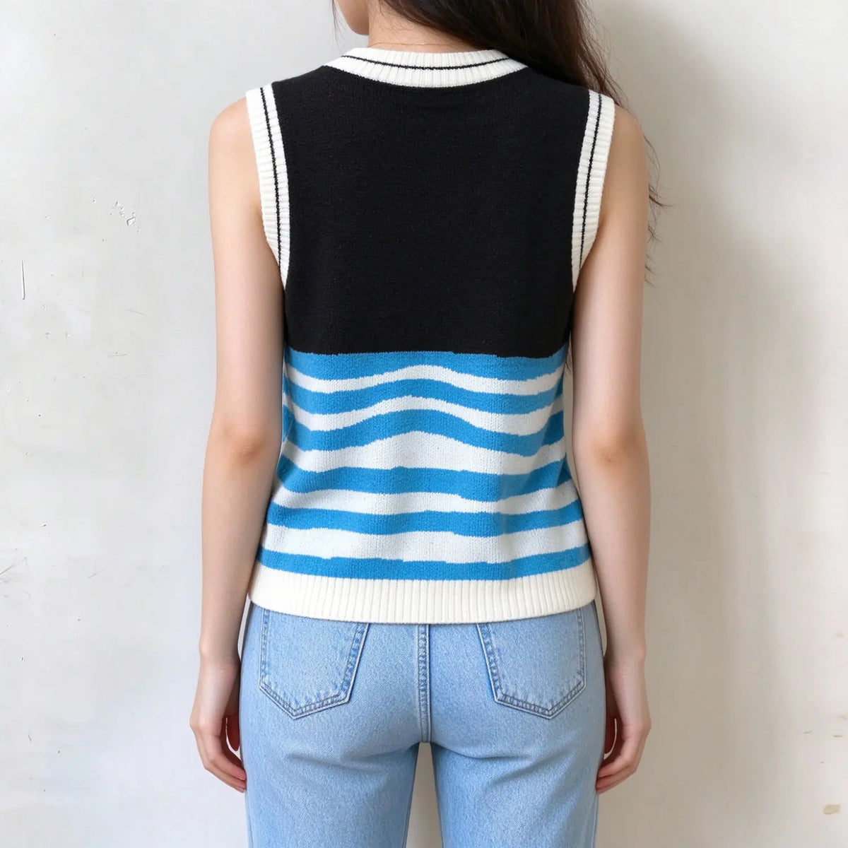 Back view of female model wearing sleeveless knit vest, solid black upper back, blue and white wavy striped lower back, white ribbed neck and arm trim