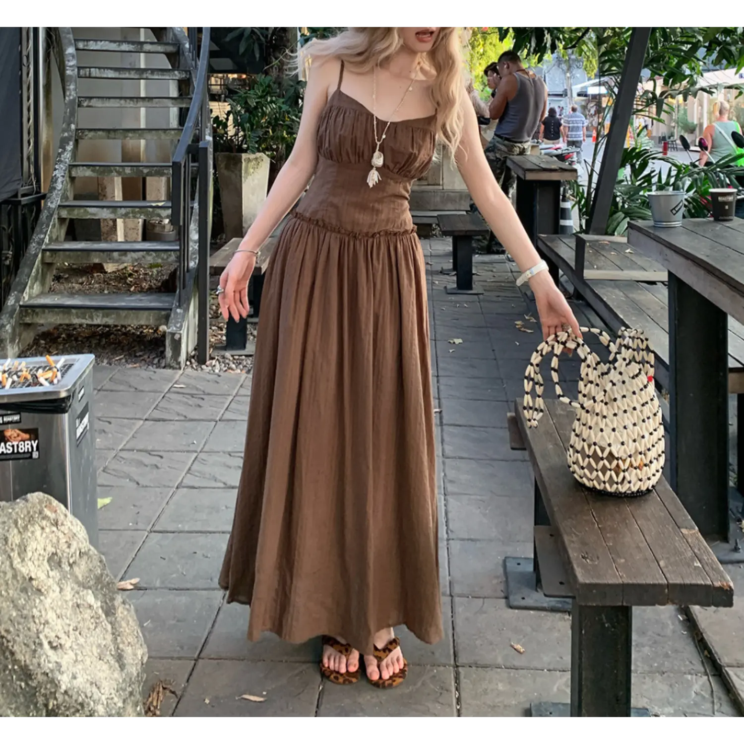 Outdoor café sidewalk full shot of mocha brown textured cami maxi dress, thin adjustable spaghetti straps, ruched sweetheart bust, ruffled empire waist seam, full flowy gathered A-line floor skirt, lightweight wrinkle fabric, styled with leopard print slide sandals, rattan woven handbag, tropical street greenery & wooden table backdrop