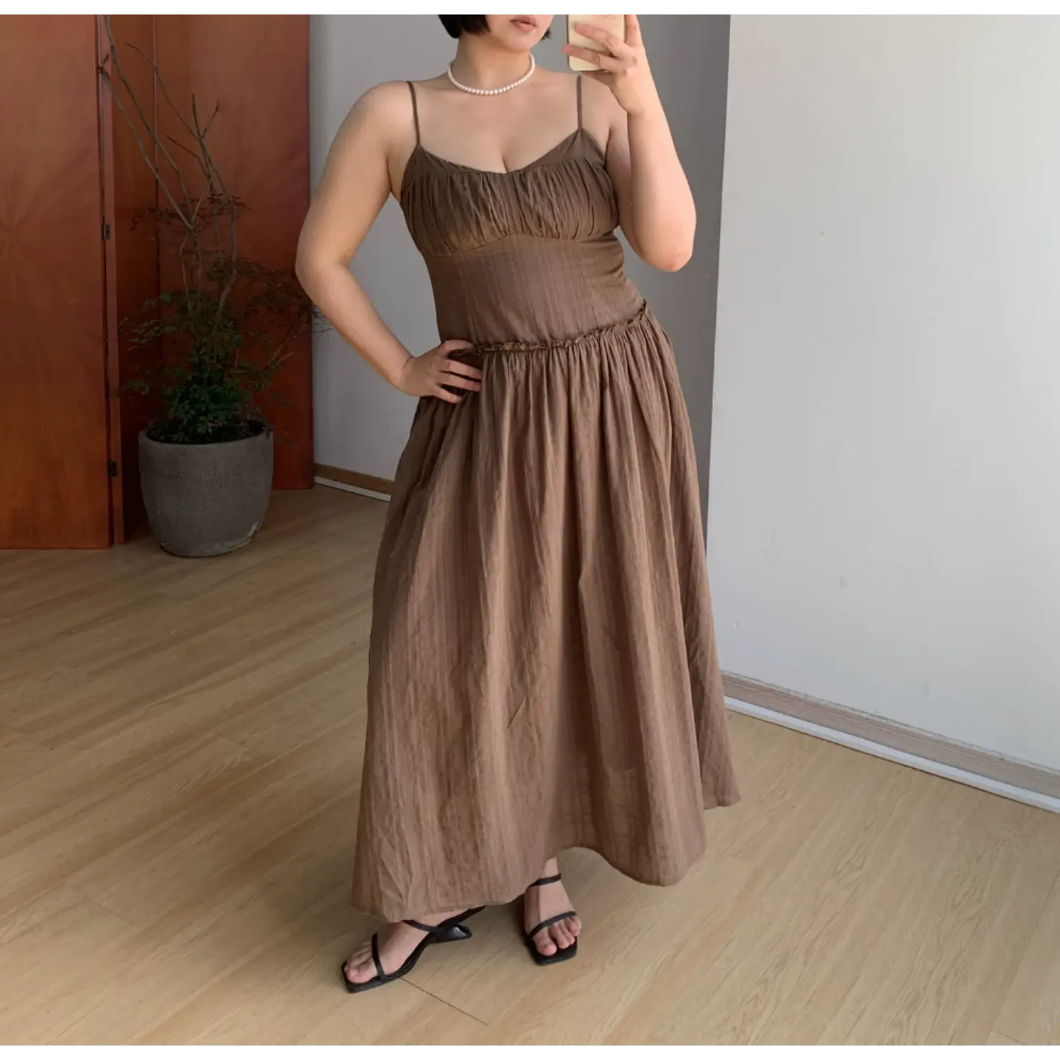 Plain front indoor mirror full shot of unlayered mocha brown slip maxi dress, thin matching spaghetti straps, ruched sweetheart V bust, tiny ruffle empire waist, heavy gathered flowy floor-length skirt, lightweight breathable textured fabric, black square strap slide sandals, simple potted plant room backdrop