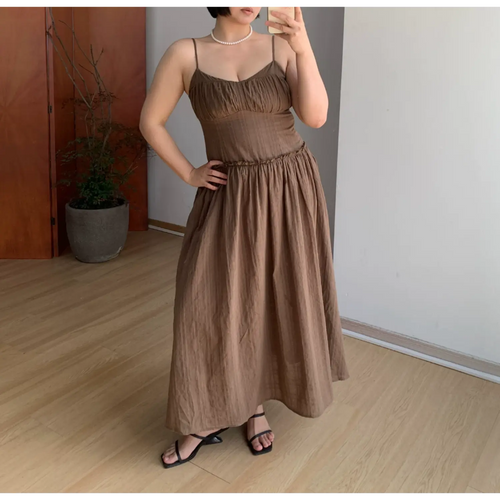 Plain front indoor mirror full shot of unlayered mocha brown slip maxi dress, thin matching spaghetti straps, ruched sweetheart V bust, tiny ruffle empire waist, heavy gathered flowy floor-length skirt, lightweight breathable textured fabric, black square strap slide sandals, simple potted plant room backdrop