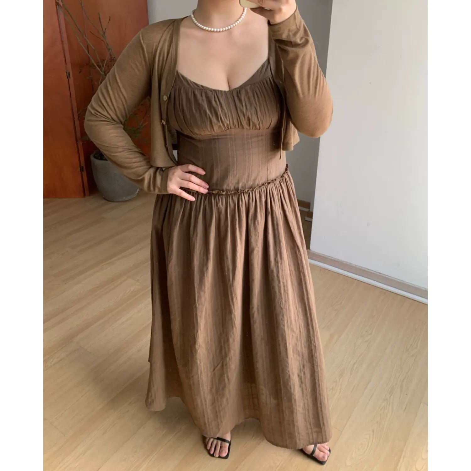Full indoor mirror layered styling shot of mocha brown textured maxi slip dress paired with matching thin long sleeve cropped knit cardigan, ruched sweetheart bust, delicate ruffle empire waist trim, full gathered flowy floor skirt, thin spaghetti straps, black thin strap slide sandals, minimalist light wood floor & potted plant decor