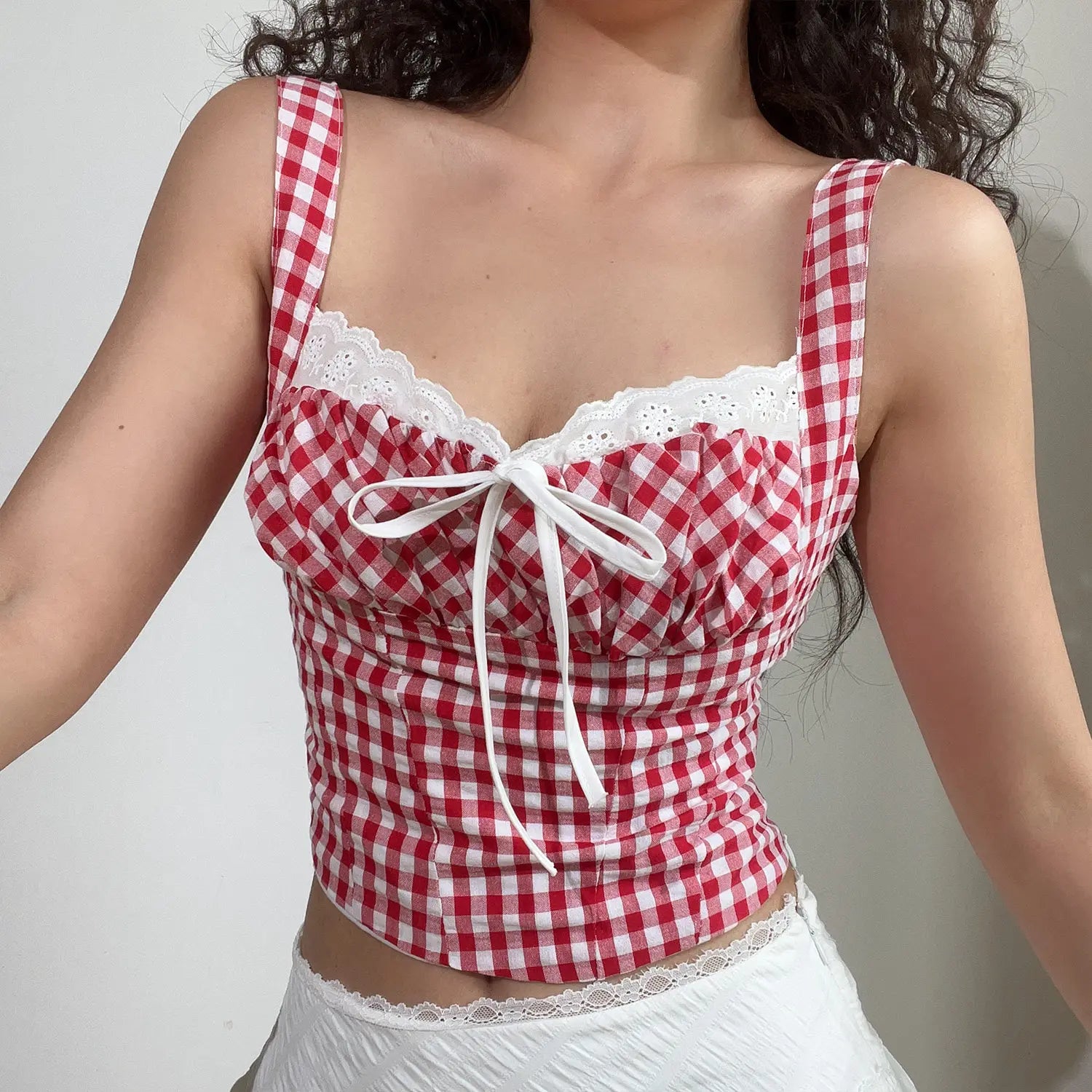 Medium close lifestyle shot of red gingham cami top with eyelet lace bust, front white ribbon tie, matching white lace hem mini skirt on model
