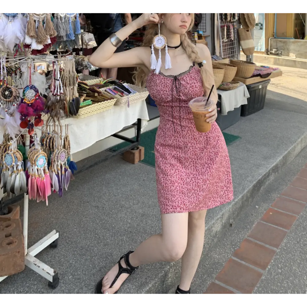 Three-quarter outdoor market lifestyle shot of pink tiny floral spaghetti strap mini dress, model grabbing white tassel dreamcatcher accessory, wearing black strappy wrap sandals