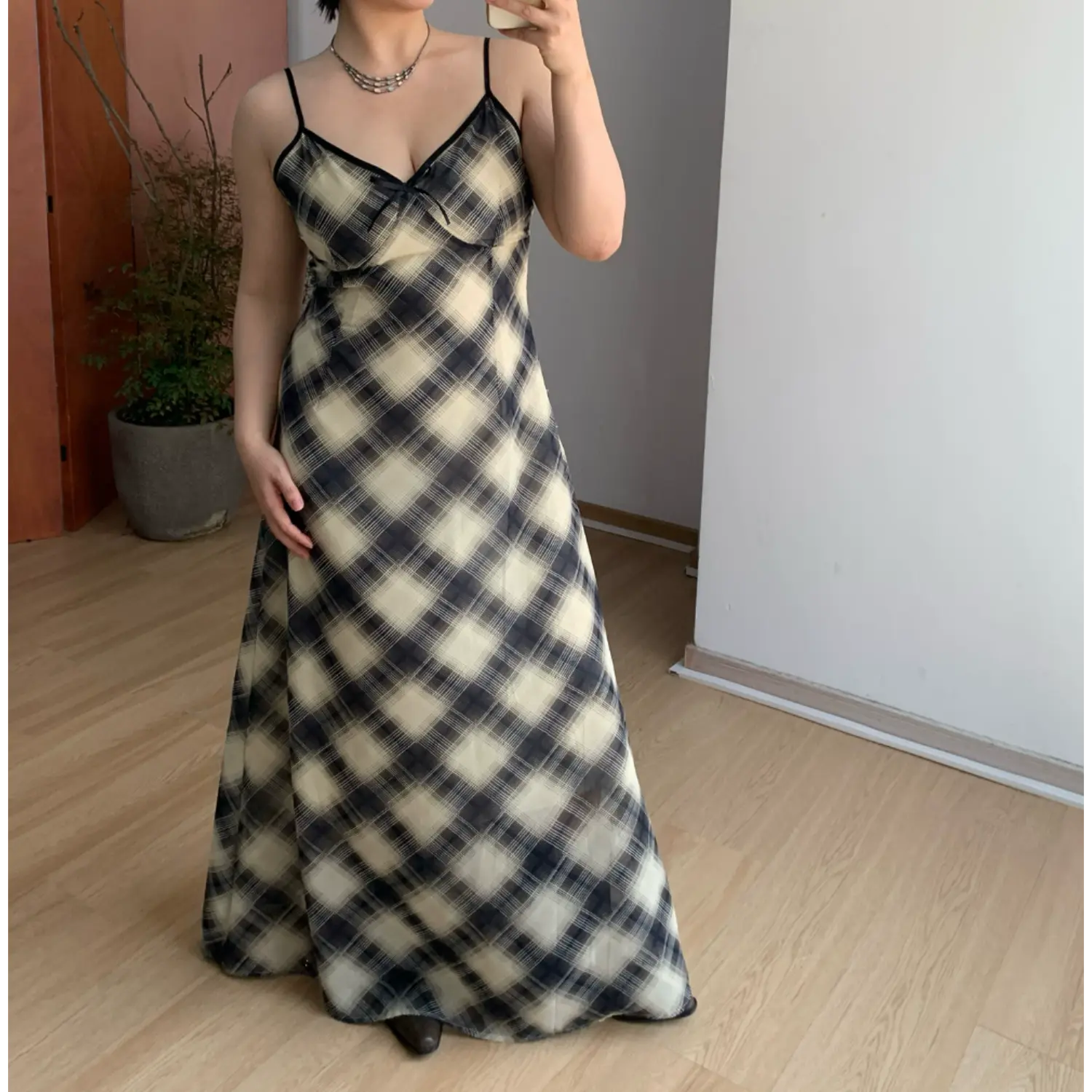 Plain front indoor mirror full shot of lightweight sheer plaid maxi slip dress, thin spaghetti straps with black contrast binding, V-neck mini fabric bow, bias cut flowy floor-length skirt, soft sheer chiffon texture, neutral minimalist room background