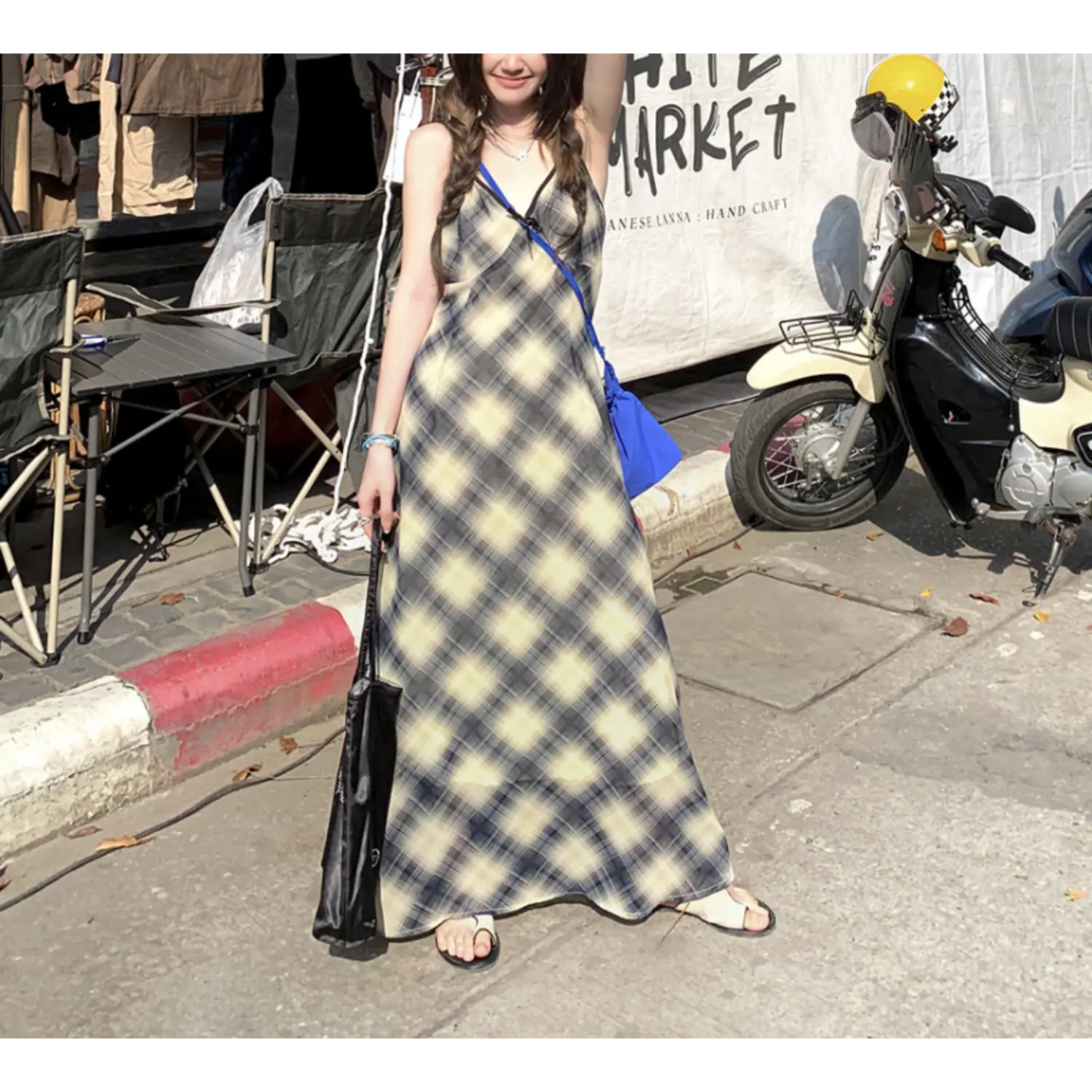 Static outdoor market full shot of lightweight sheer plaid slip maxi dress, V-neck cami design with tiny front bow, flowy floor skirt, styled with white slide sandals, holding black printed canvas tote bag, vintage scooter & handcraft market banner backdrop