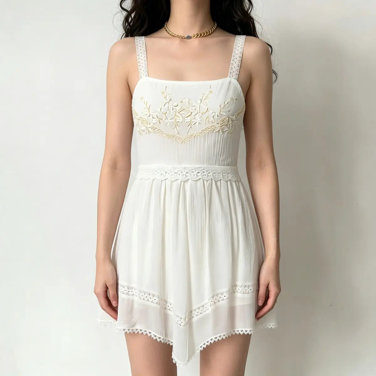 Model wearing a cream sundress with gold floral embroidery, lace-trimmed spaghetti straps, and an asymmetrical lace hem.