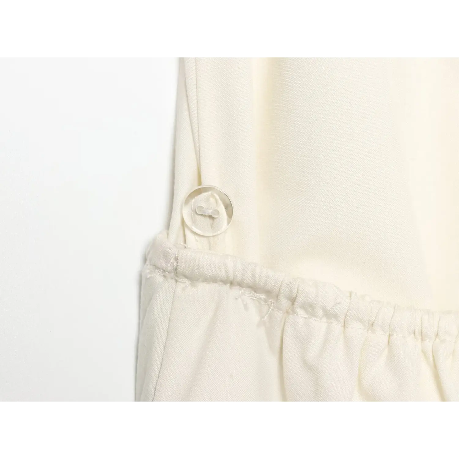 Close‑up of side hidden button fastener at elastic waist seam on cream halter sundress, fine fabric stitching detail