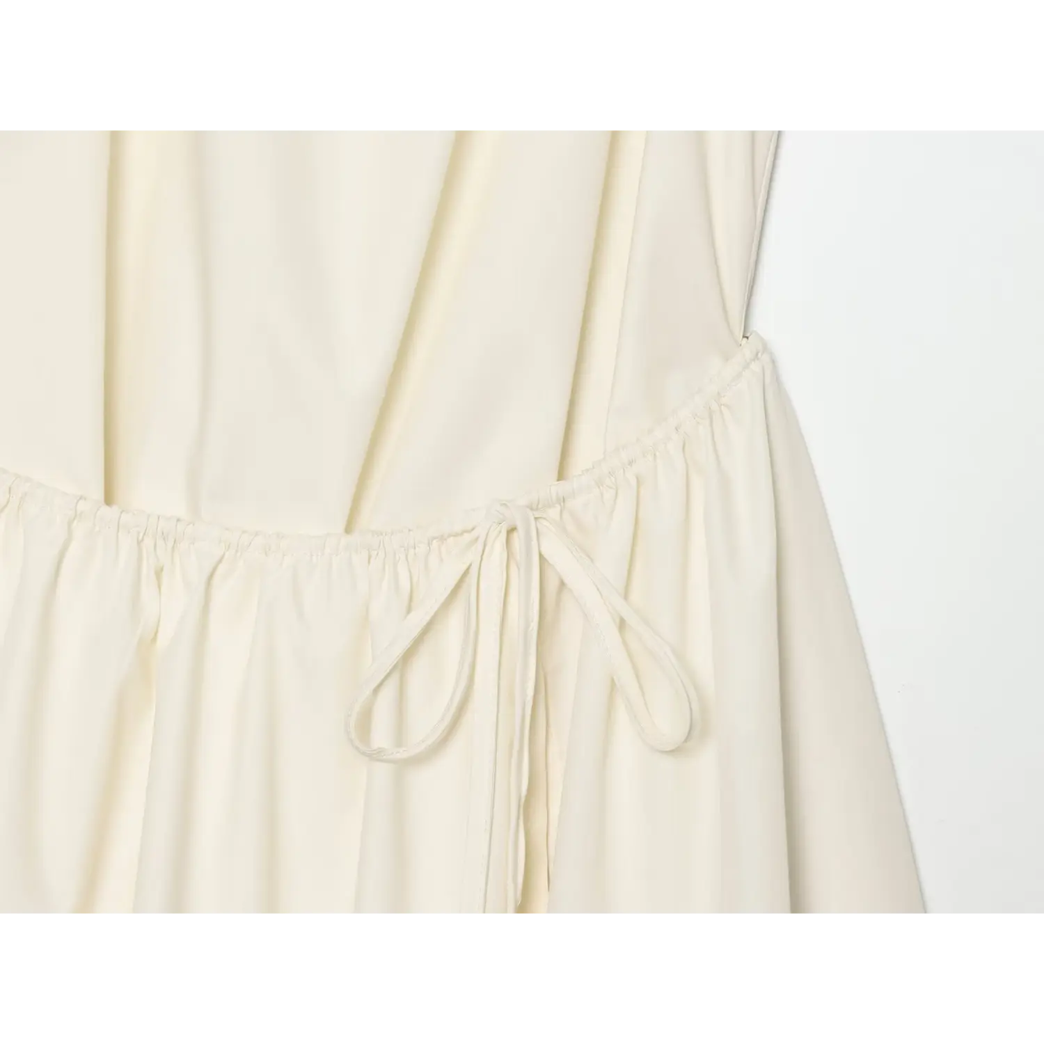 Close‑up view of side waist drawstring tie and elastic gathered seam on cream halter tiered mini dress, white background product detail shot